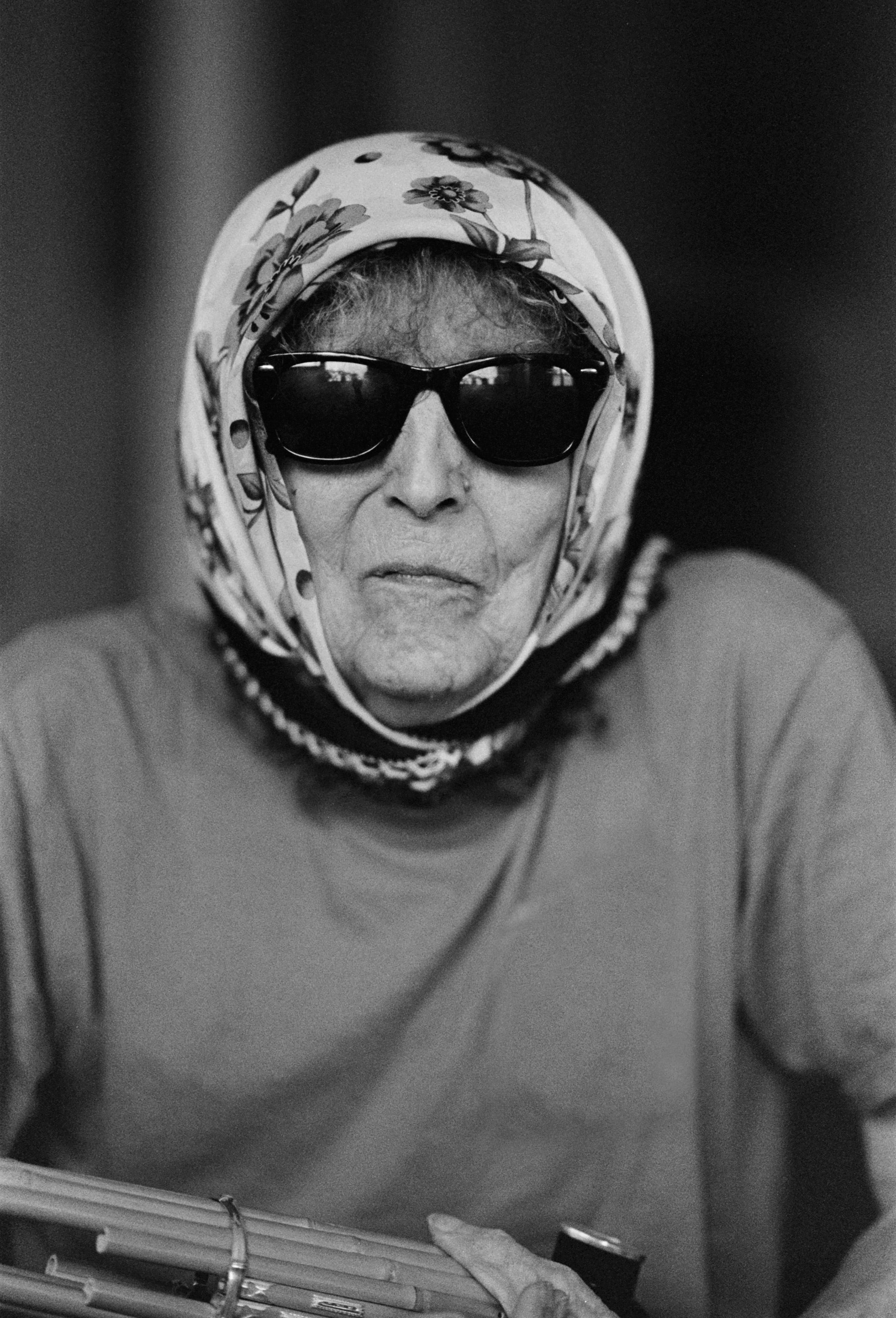 An elderly person wearing sunglasses and a floral headscarf holds an object in their hands.