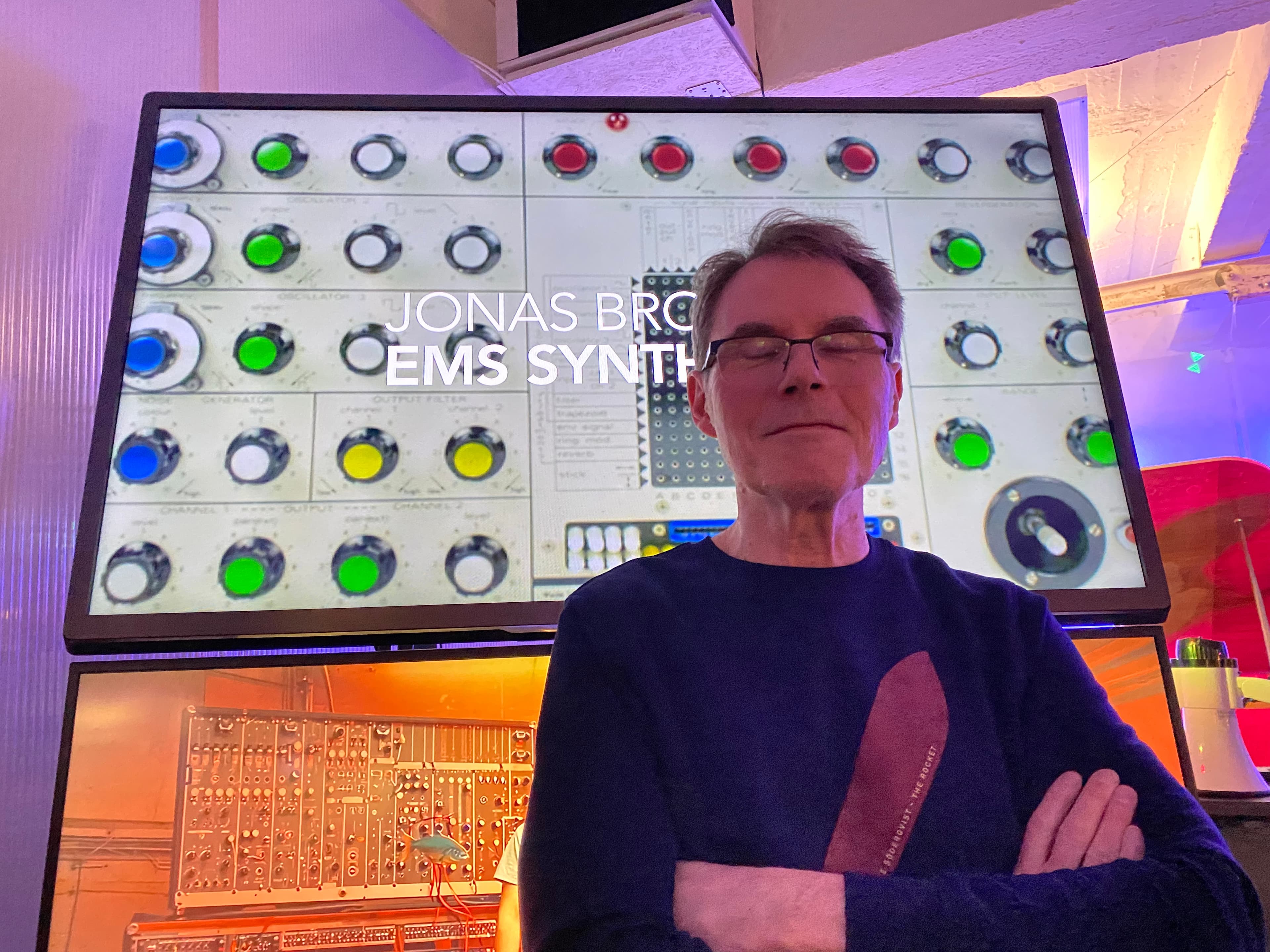 A person with glasses stands in front of a display showing a synthesizer interface labeled "Jonas Bro EMS Synth."