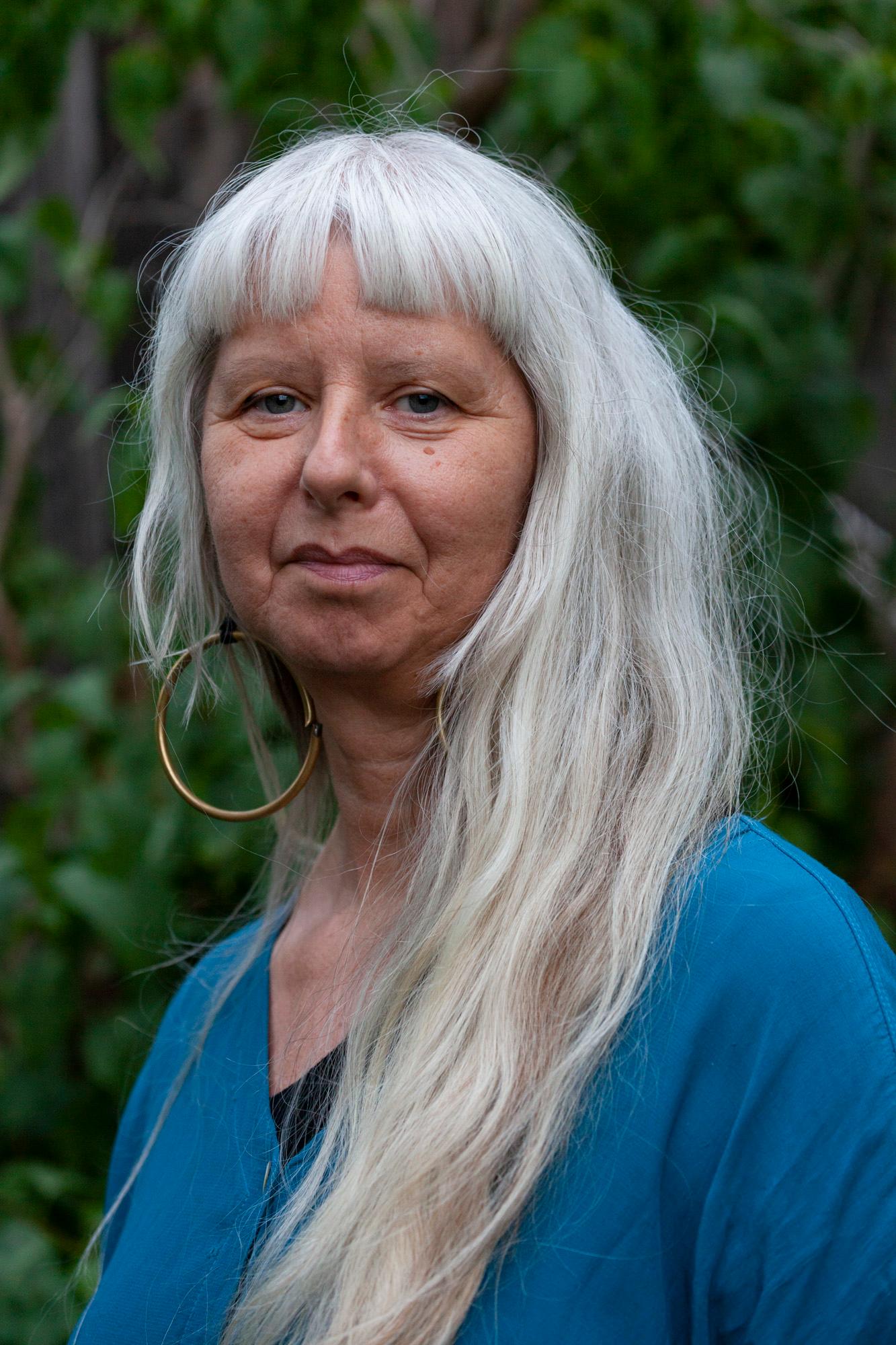 A person with long gray hair, wearing a blue top and a large hoop earring, is standing in front of a leafy background.