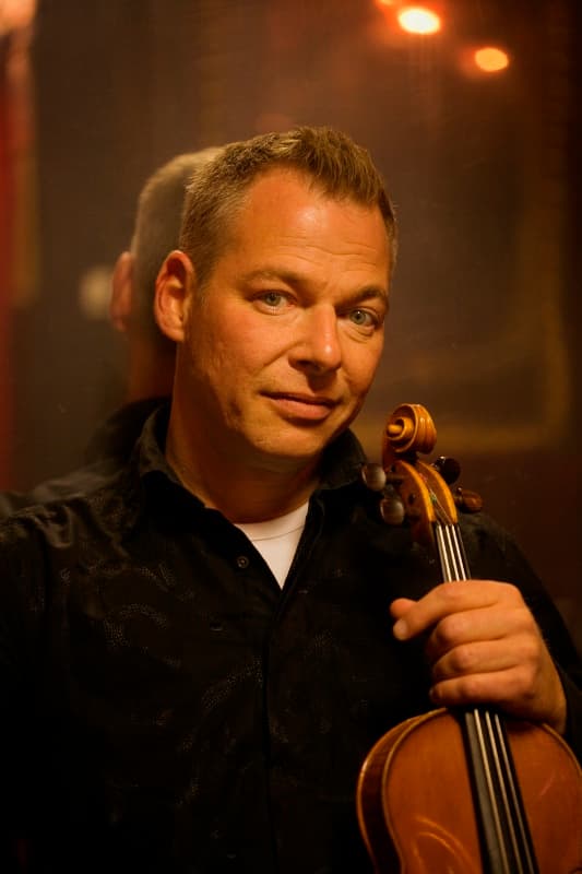 A man holding a violin, looking at the camera, with a reflective background and warm lighting.