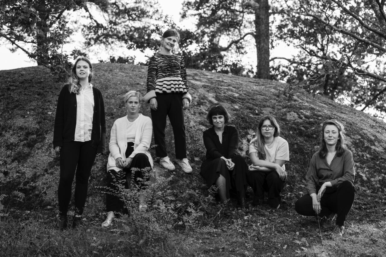 Six women are gathered outdoors on a grassy and rocky area with trees in the background, with two standing and four seated or squatting.