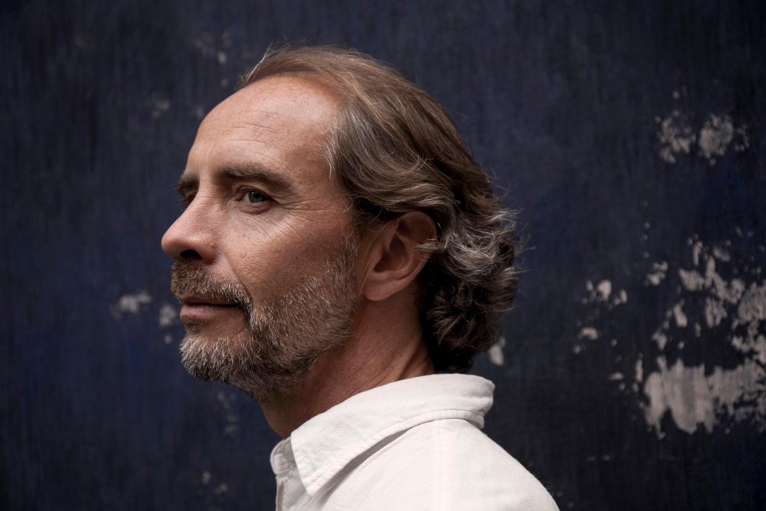 A man with gray hair and a beard is shown in profile against a dark, textured background.