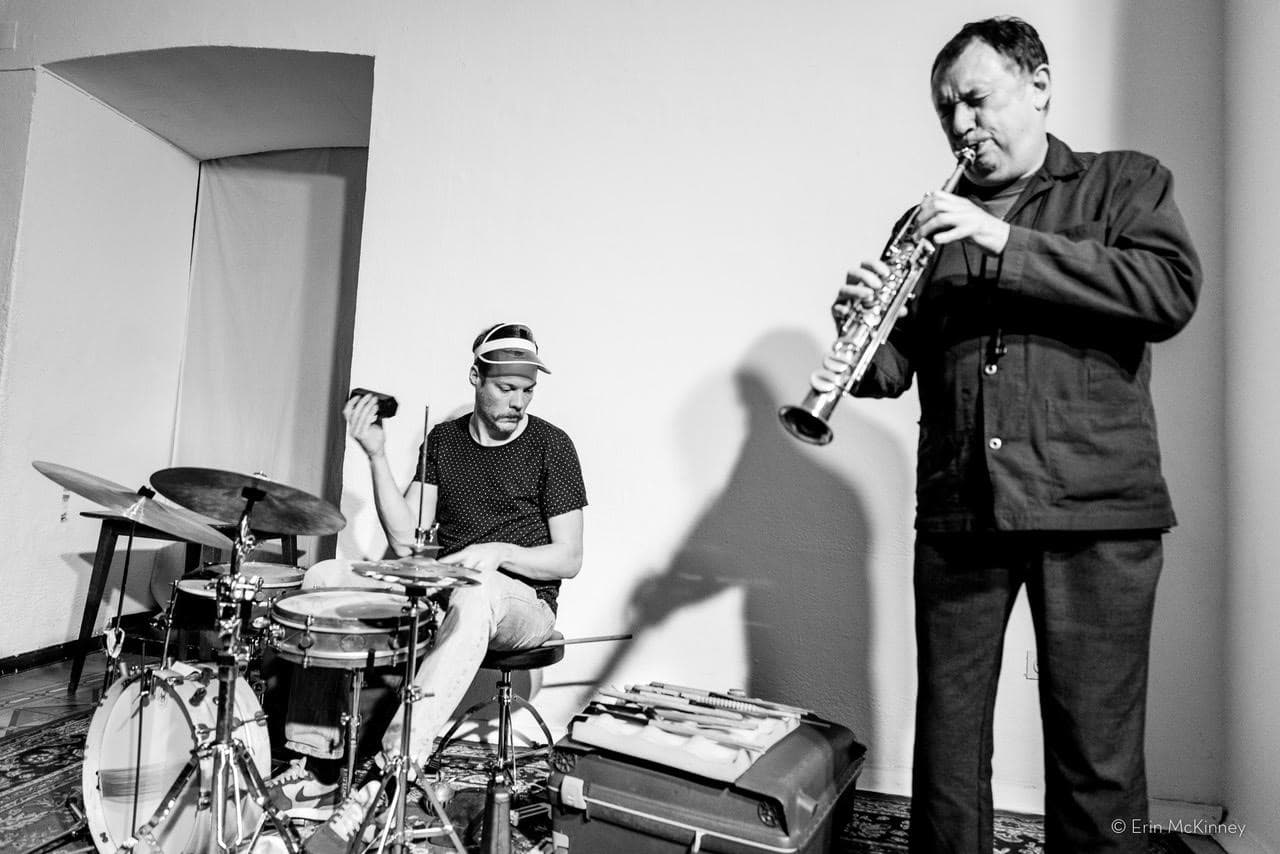 A drummer and a saxophonist perform together in a room, with the drummer seated and the saxophonist standing.