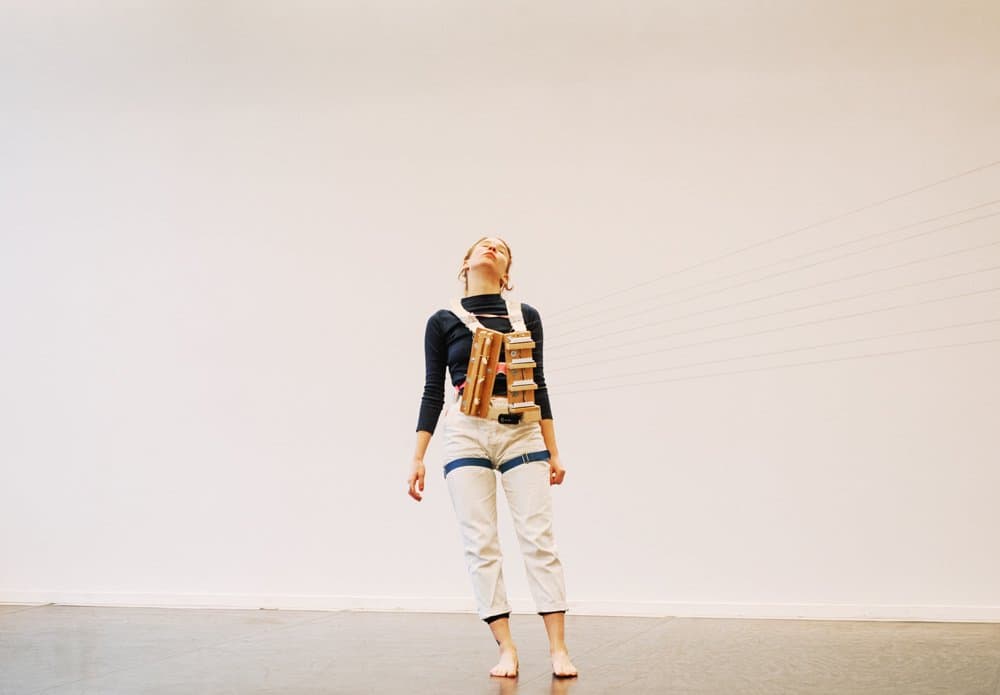 A person stands barefoot in an indoor space, wearing a device with multiple rods attached to their torso and head.