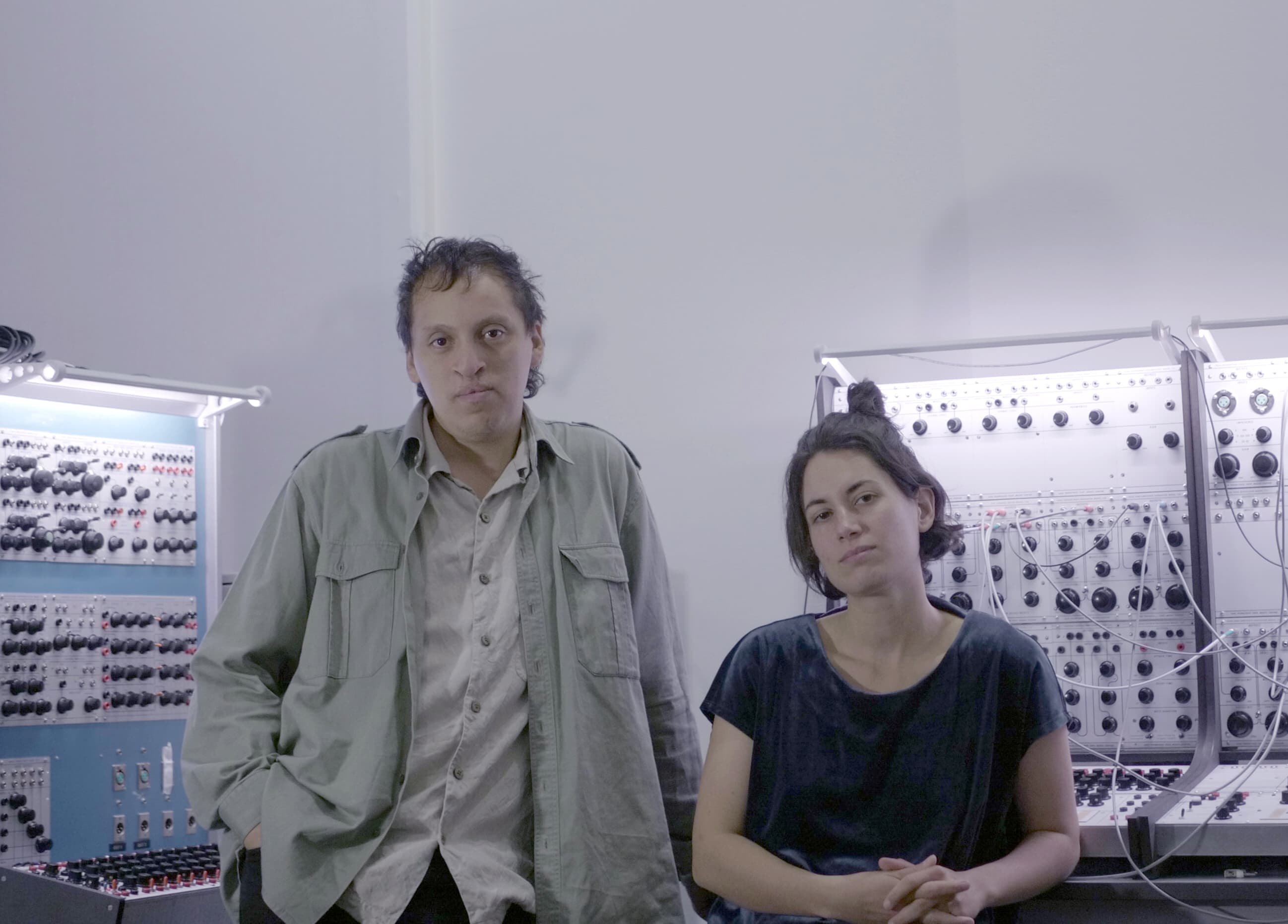 Two people are standing in front of a modular synthesizer setup in a studio.