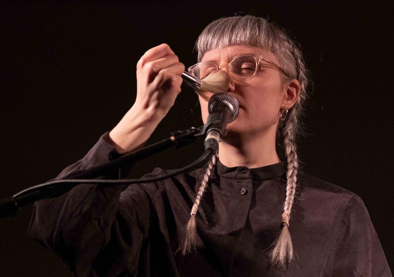 A person with braided hair and glasses holds a brush near a microphone, appearing to perform or create sound.