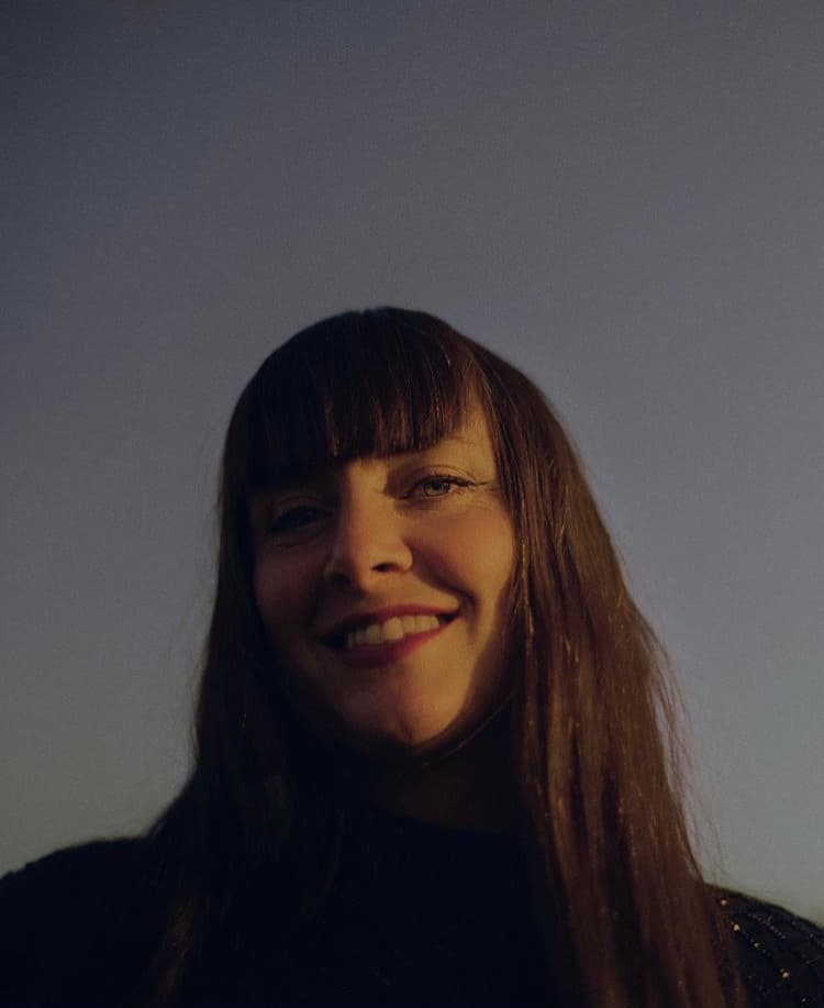 A woman with long hair and bangs smiles while looking upwards against a clear sky background.