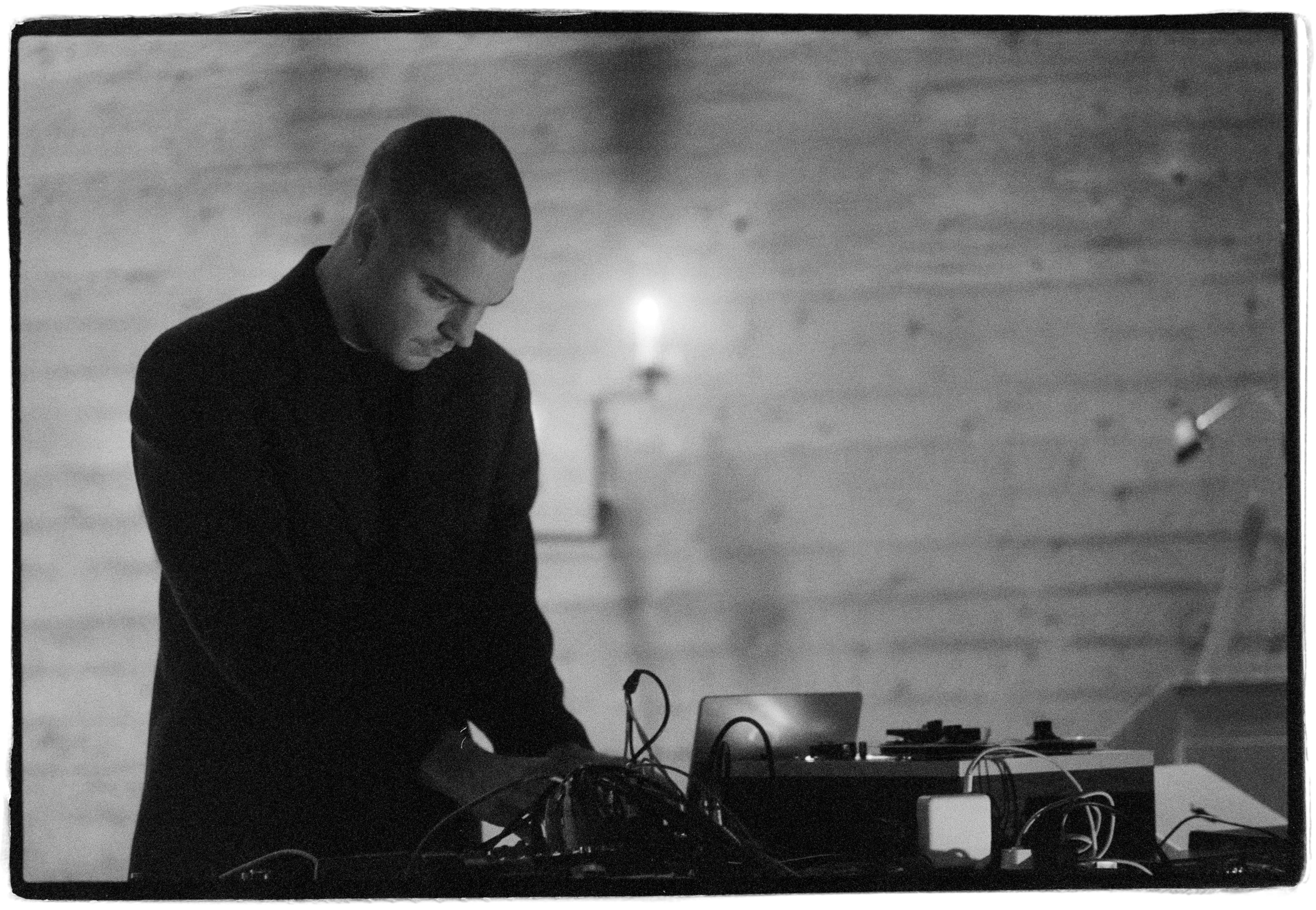 A person in a dark suit operates electronic equipment, with a blurred background featuring a candle on a wall.
