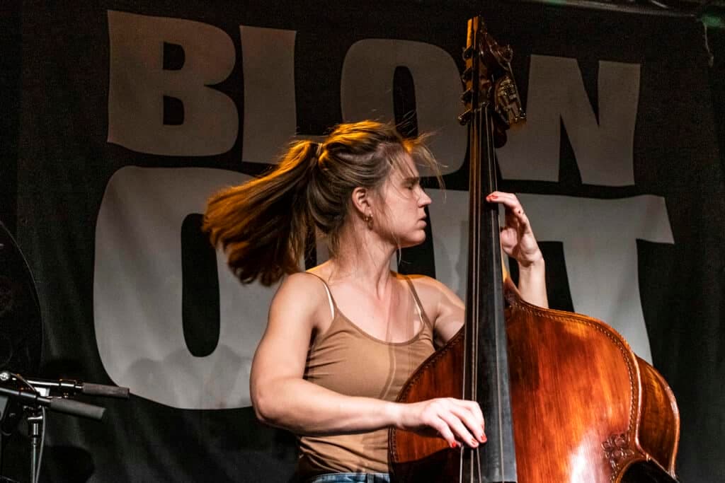 A woman is playing a double bass on stage with a "BLOW OUT" banner in the background.