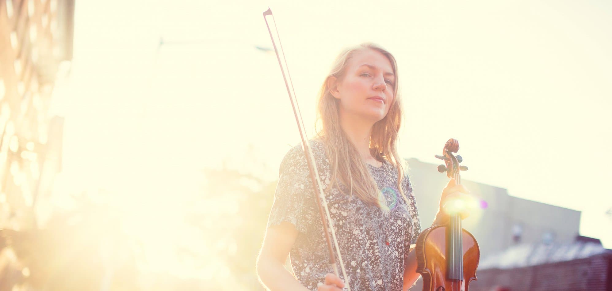 A person with long hair is holding a violin and bow, standing outside with bright sunlight in the background.