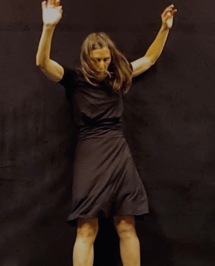 A woman in a black dress poses with arms raised against a dark background.