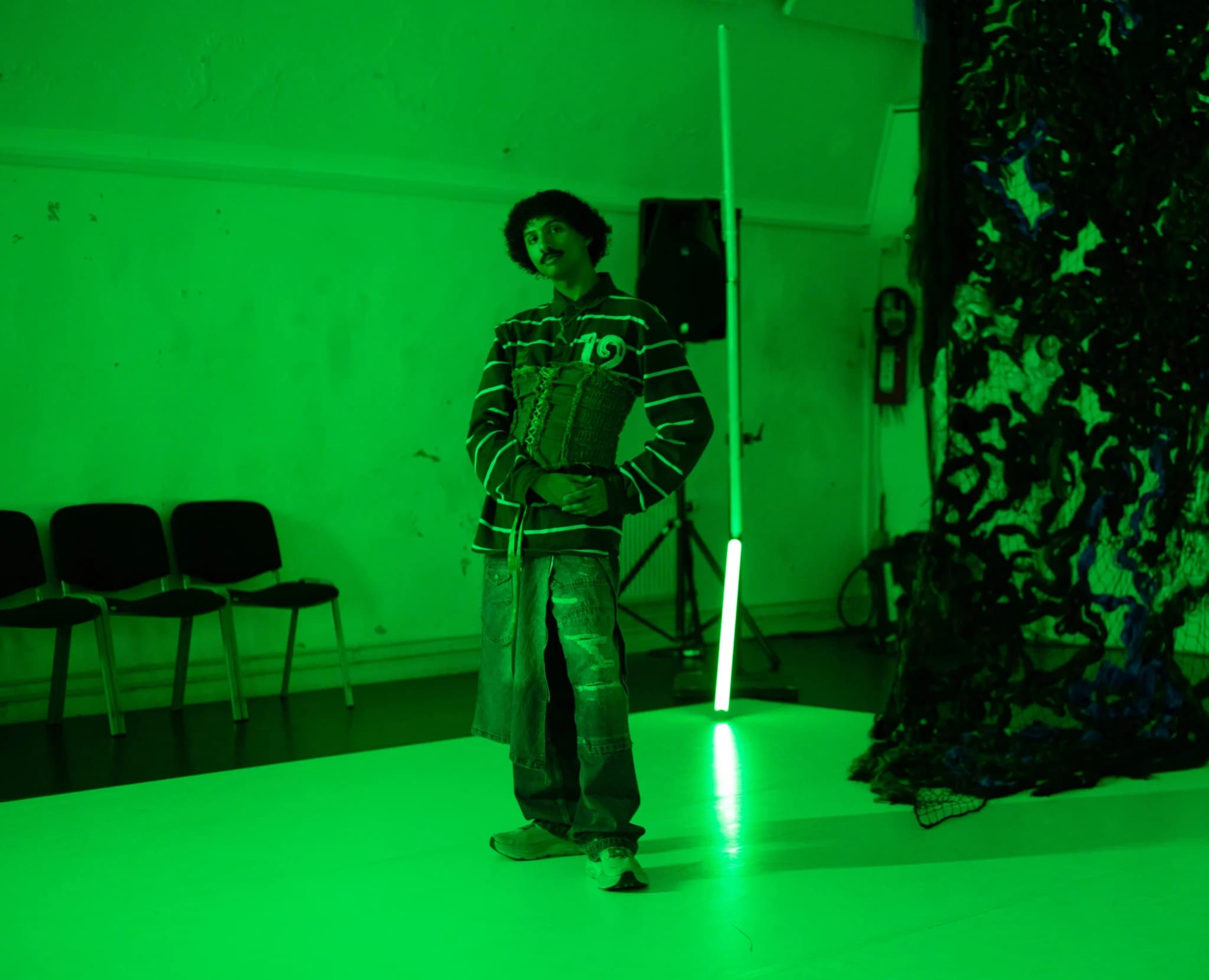 A person stands in a green-lit room, wearing a striped shirt and distressed jeans, with chairs and abstract decor in the background.