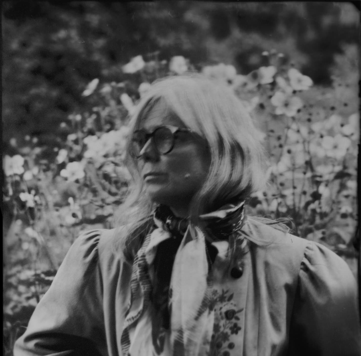 A person with long hair and glasses stands outdoors, surrounded by flowers, wearing a scarf and a jacket.