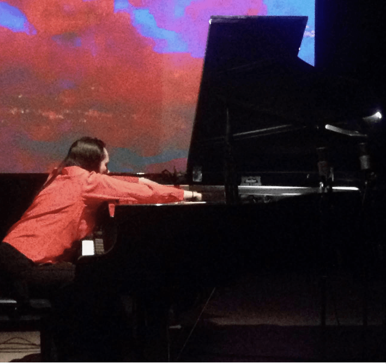 A person in a red shirt is playing a grand piano on a stage with a colorful backdrop.