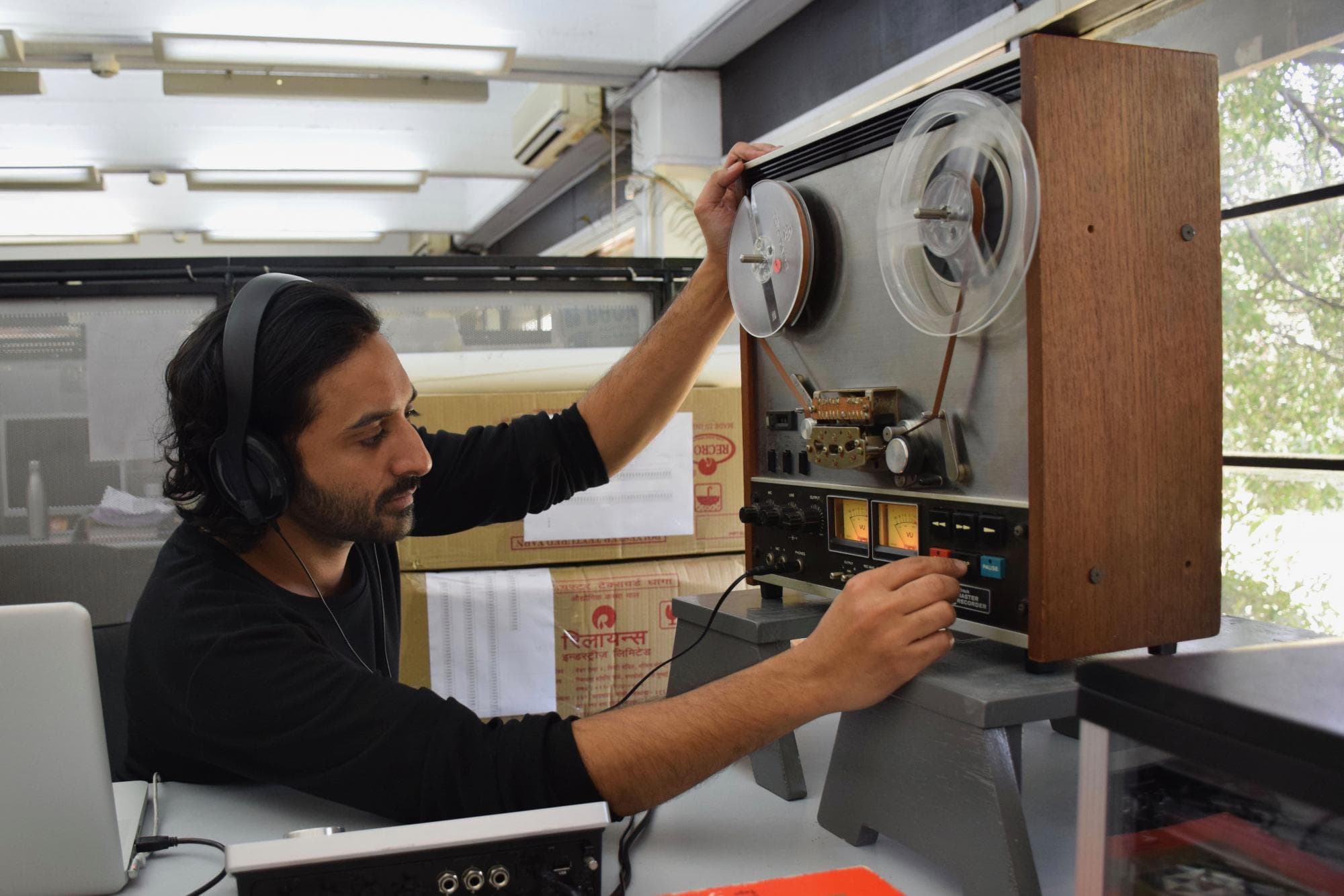 A person wearing headphones operates a reel-to-reel tape recorder in an office setting.