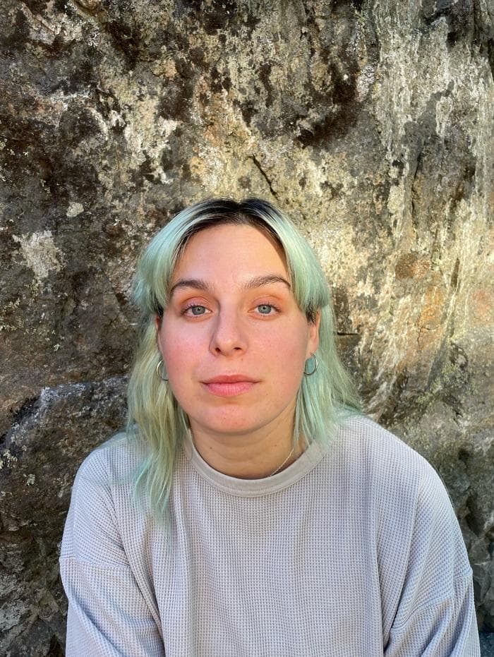 A person with light green hair is standing in front of a rocky background, wearing a light gray sweater.