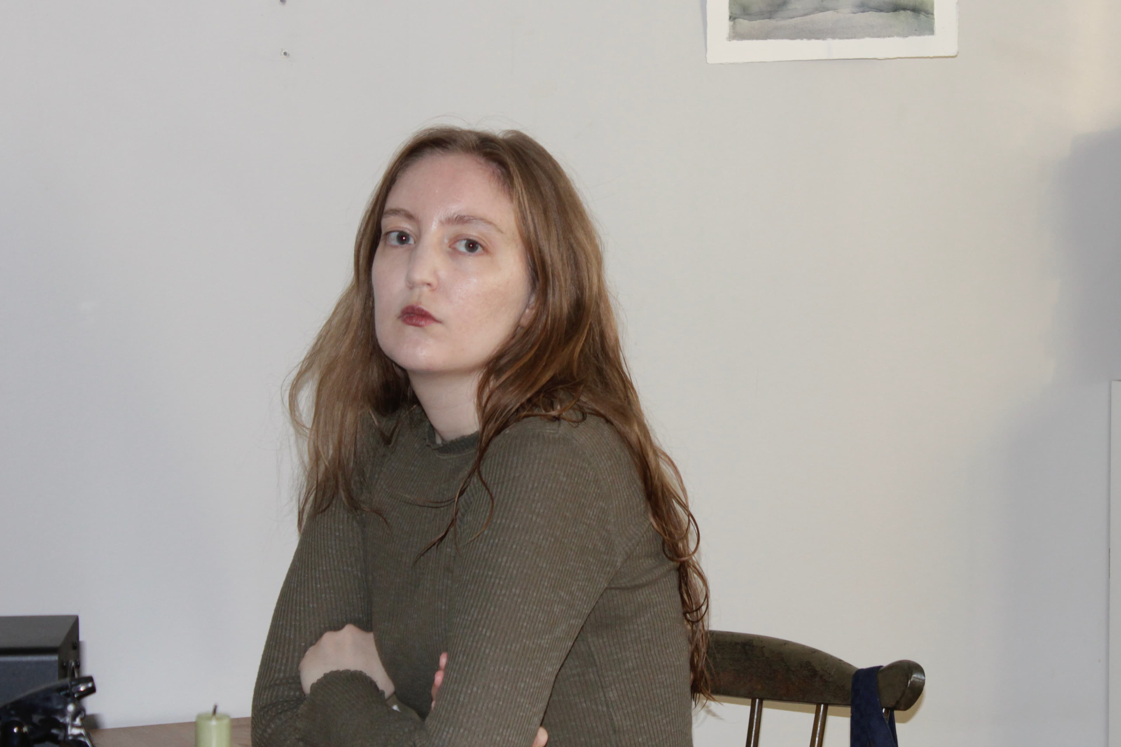 A person with long hair and a neutral expression sits at a table in a room with a light-colored wall.