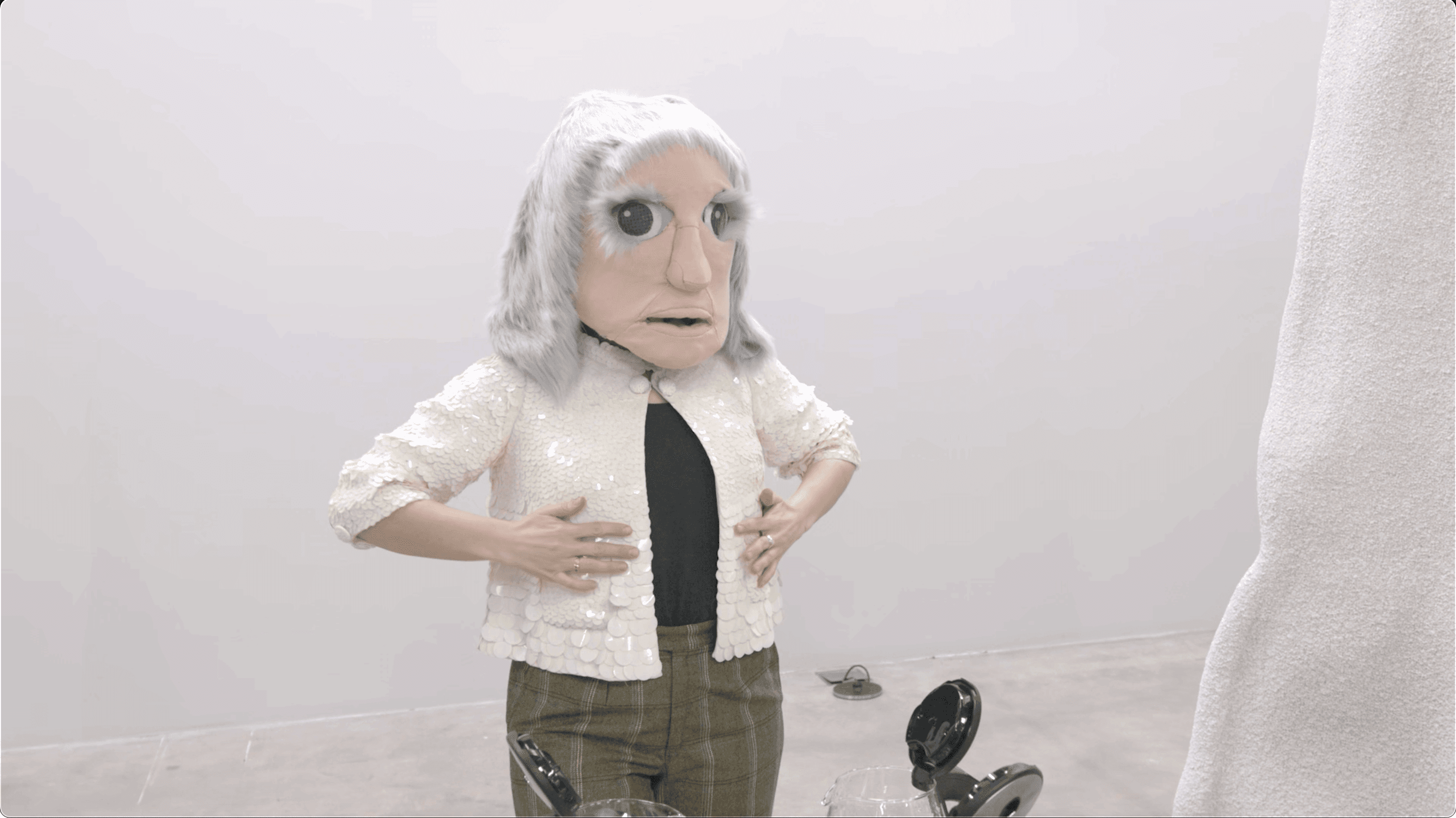 A person wearing a large mask with exaggerated facial features and a white sequined jacket, standing in a room with a gray floor.