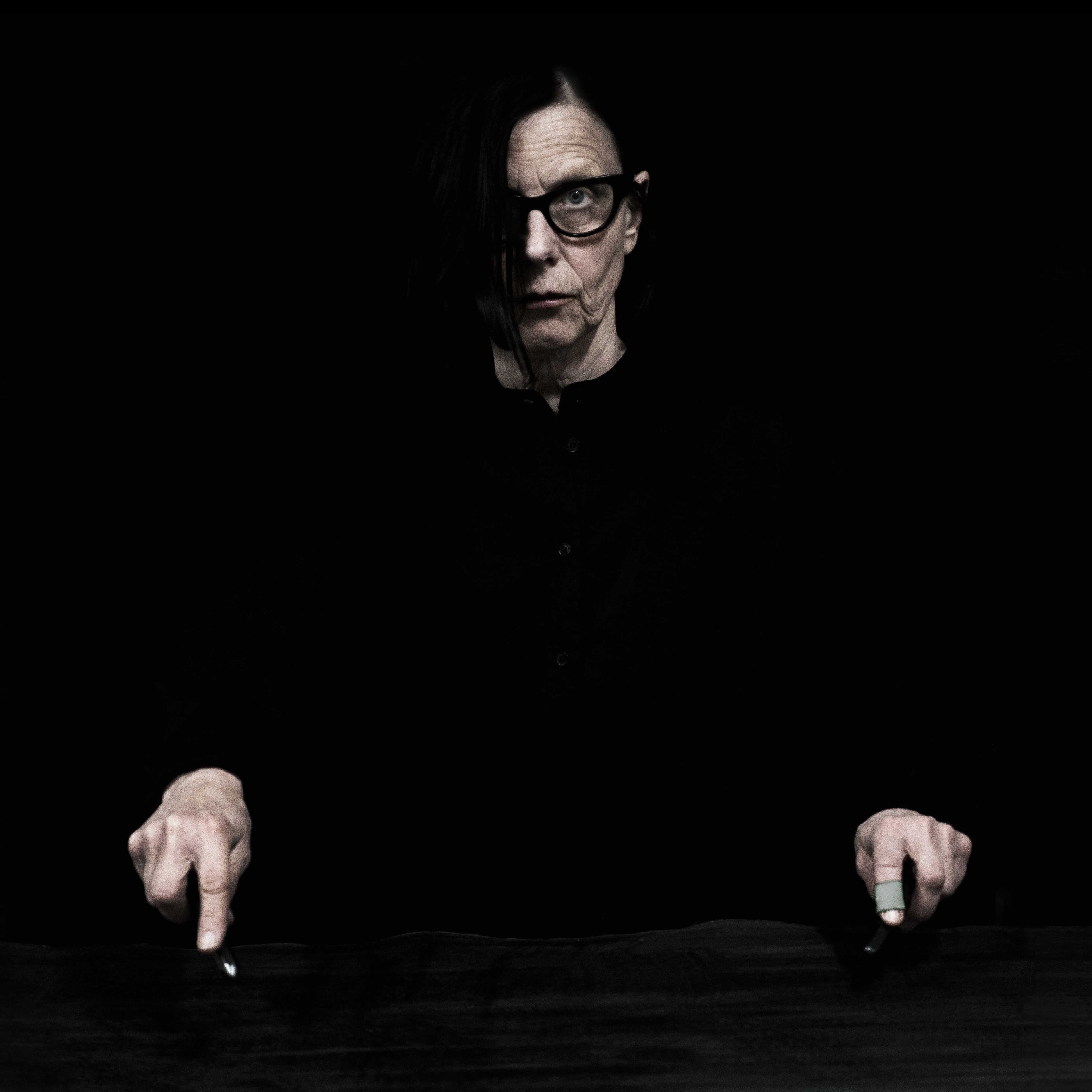 A person wearing glasses and dark clothing points downward with both hands in a dimly lit setting.