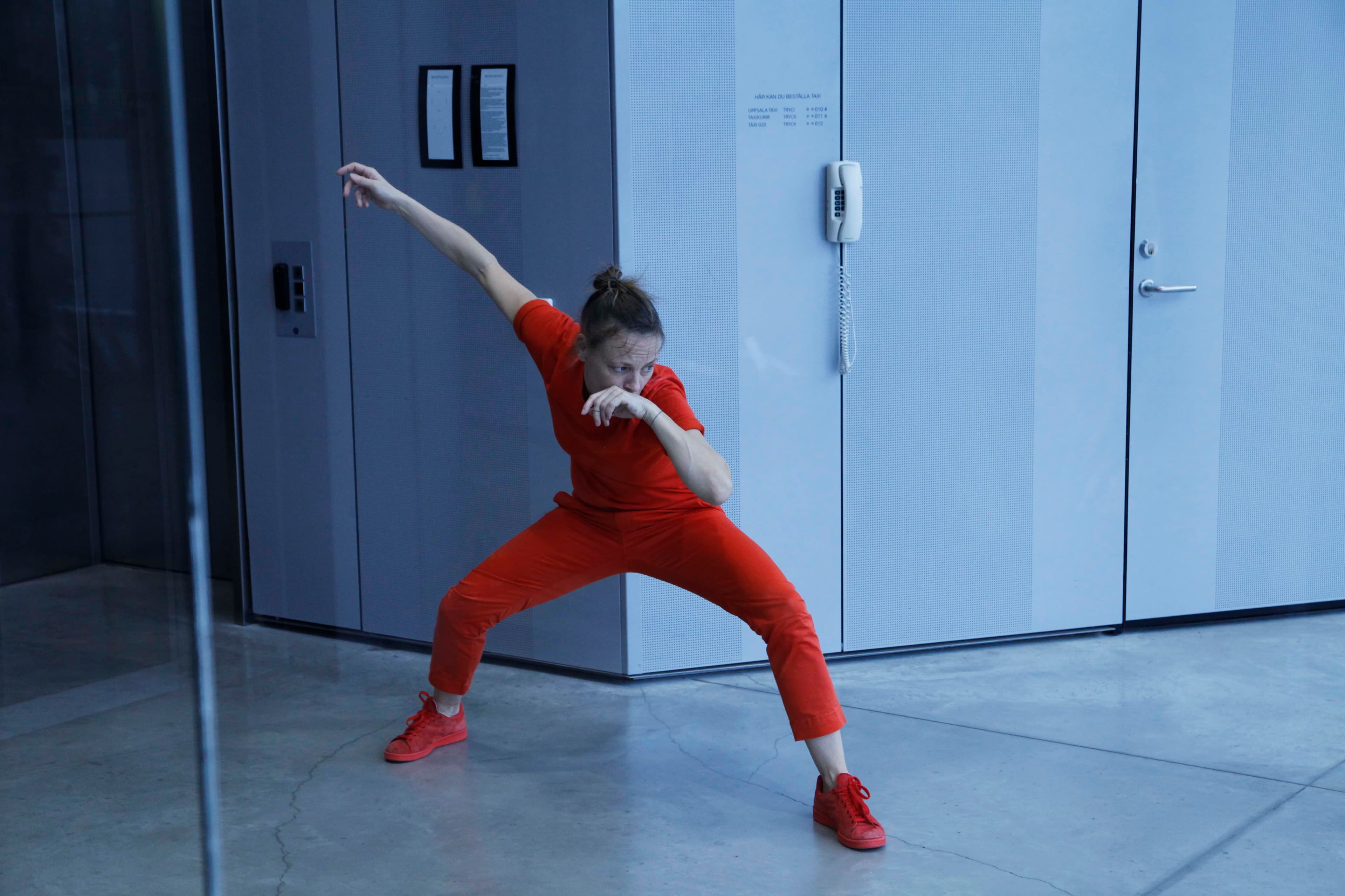 A person in a red outfit strikes a dynamic pose in a modern, minimalist room with a wall-mounted telephone.