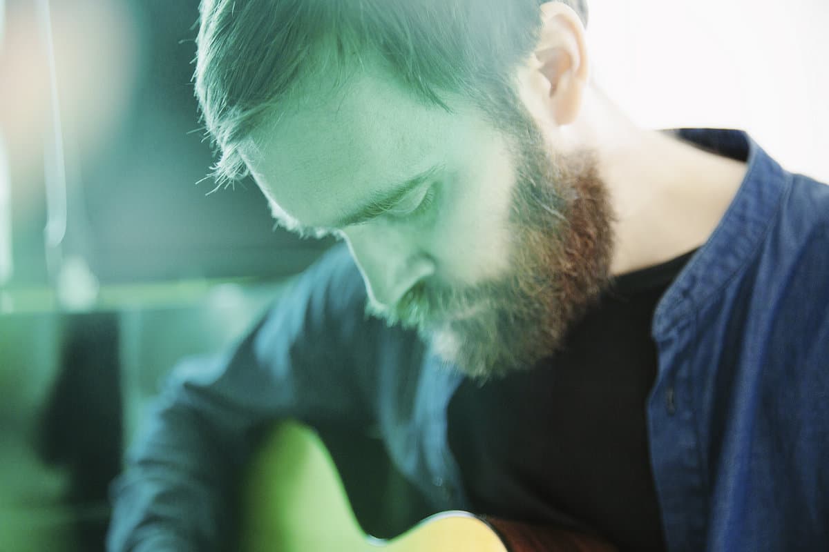 A bearded man with a downcast gaze is playing a guitar.