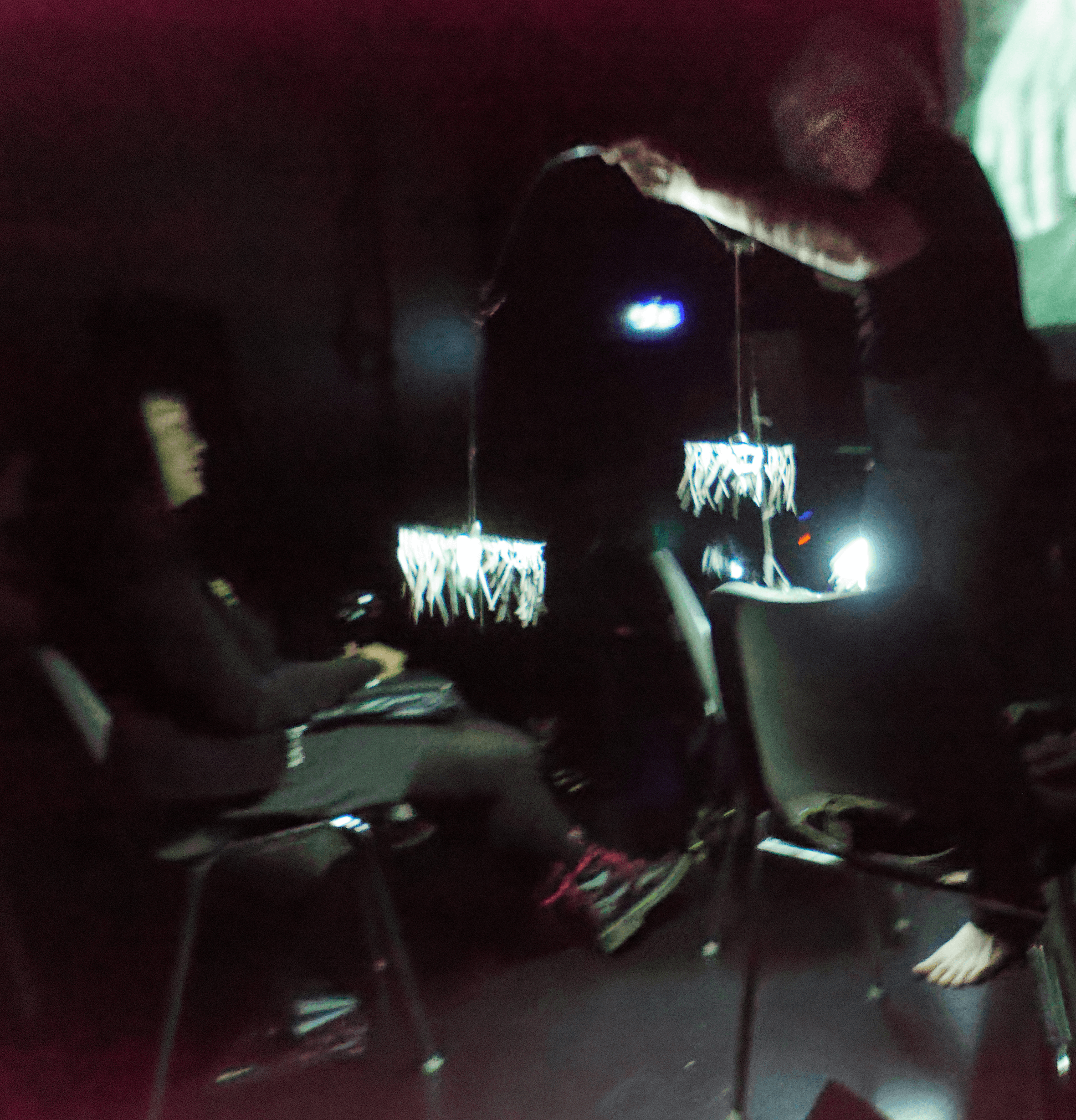 A person is holding a light fixture with hanging elements in a dimly lit room, while another person sits nearby.