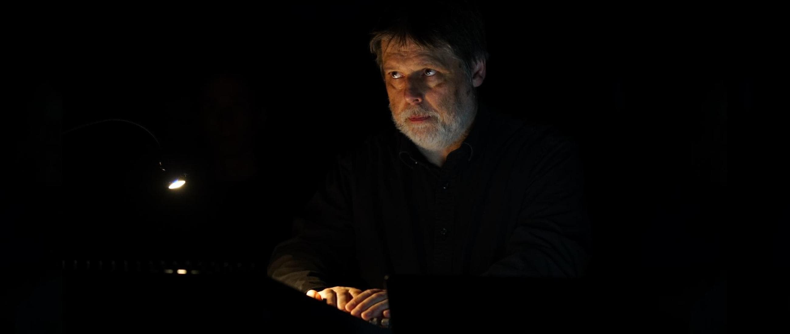 A man with a gray beard is illuminated by a small light in an otherwise dark setting, looking intently ahead.