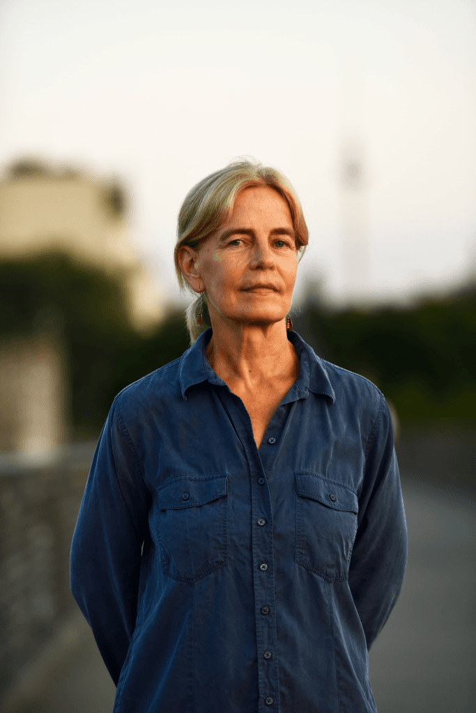 A woman with blonde hair wearing a blue button-up shirt stands outdoors, with a blurred background of greenery and buildings.