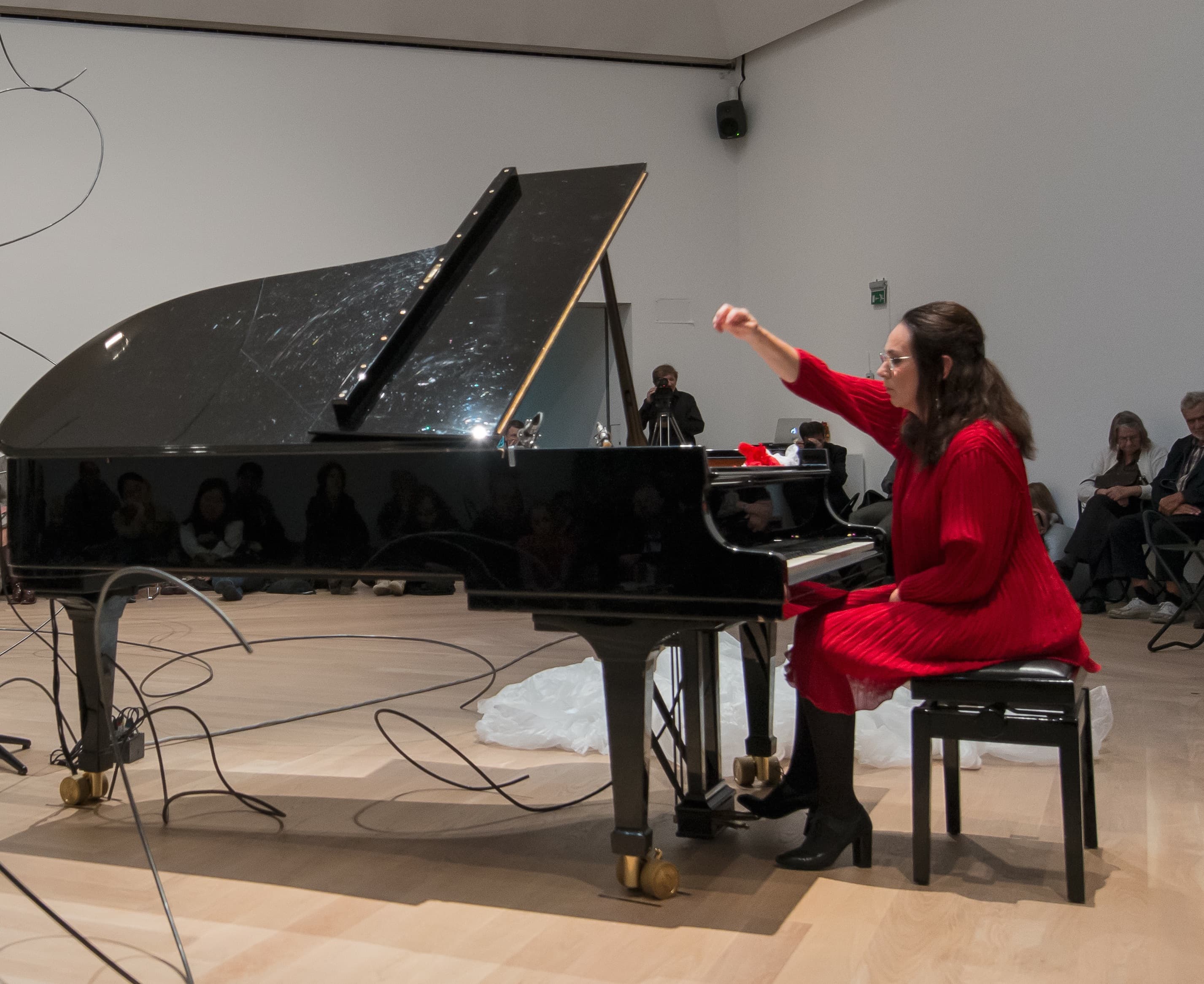 A woman in a red dress plays a grand piano in an art gallery setting, with an audience seated around her.